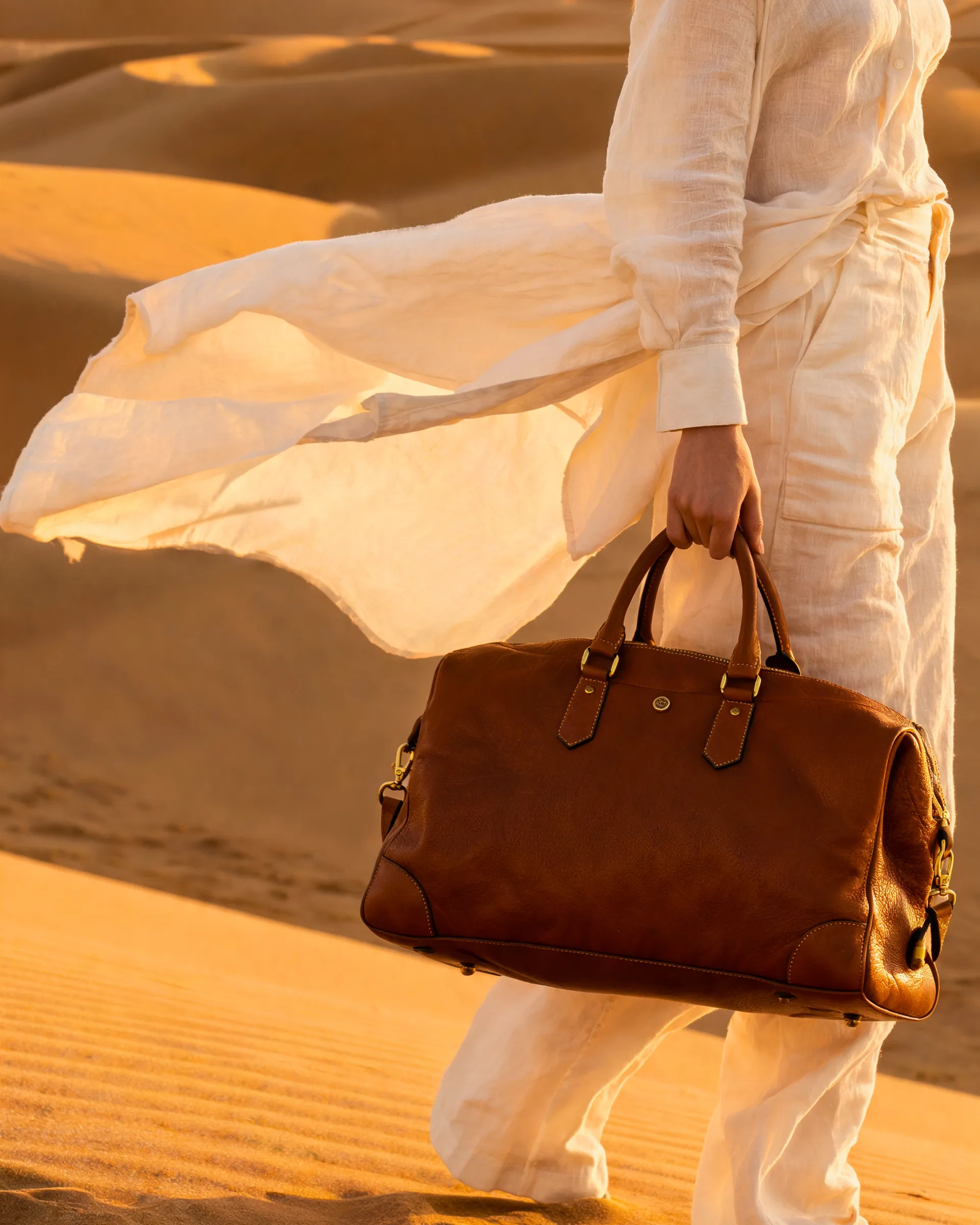 A traveler in cream linen carries a cognac leather weekender across golden dunes at last light